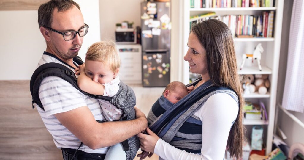 babywearing-mom-and-dad-holding-their-babies-in-carriers-smiling-at-each-other