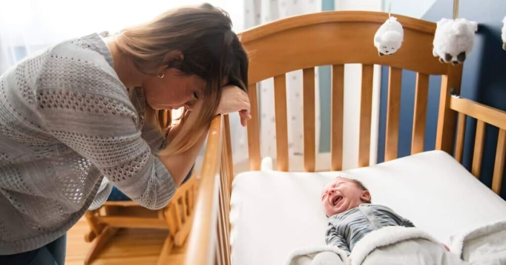 A tired mother leans on her baby's crib, looking down at her crying infant who won't sleep. The scene captures the exhaustion and frustration often felt by parents dealing with a sleepless baby.