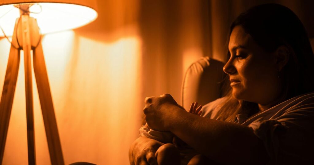 overnight-doula-for-twins-mother-sitting-on-chair-next-to-a-lamp-in-warm-lighting-at-night-holding-her-baby
