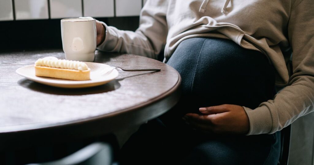 Pregnant woman sitting with a white mug in hand and food in the table