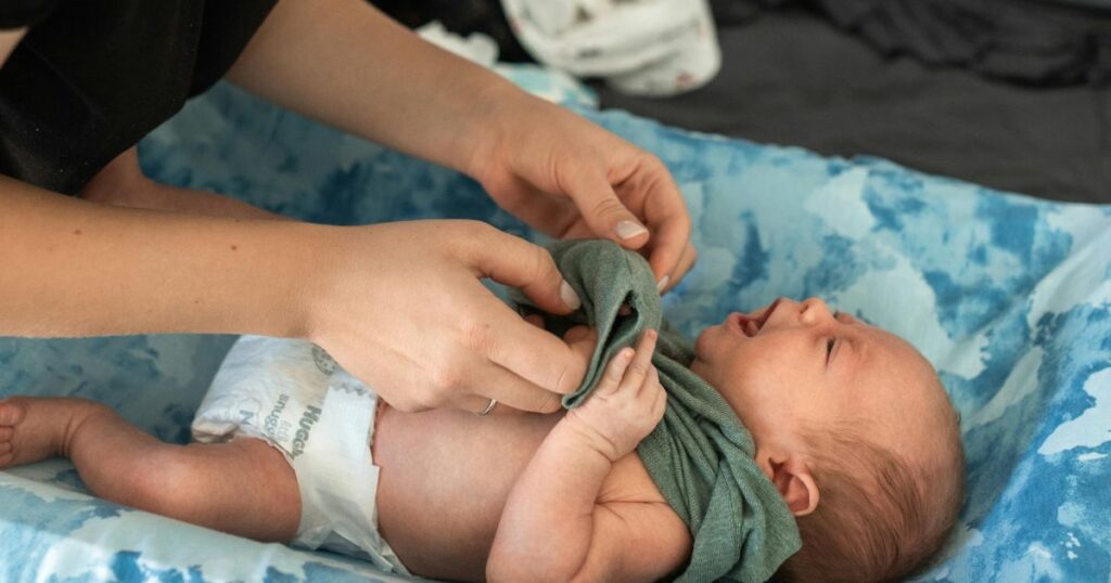 postpartum doula changing a newborn baby on a blue changing table and dressing him in a onesie