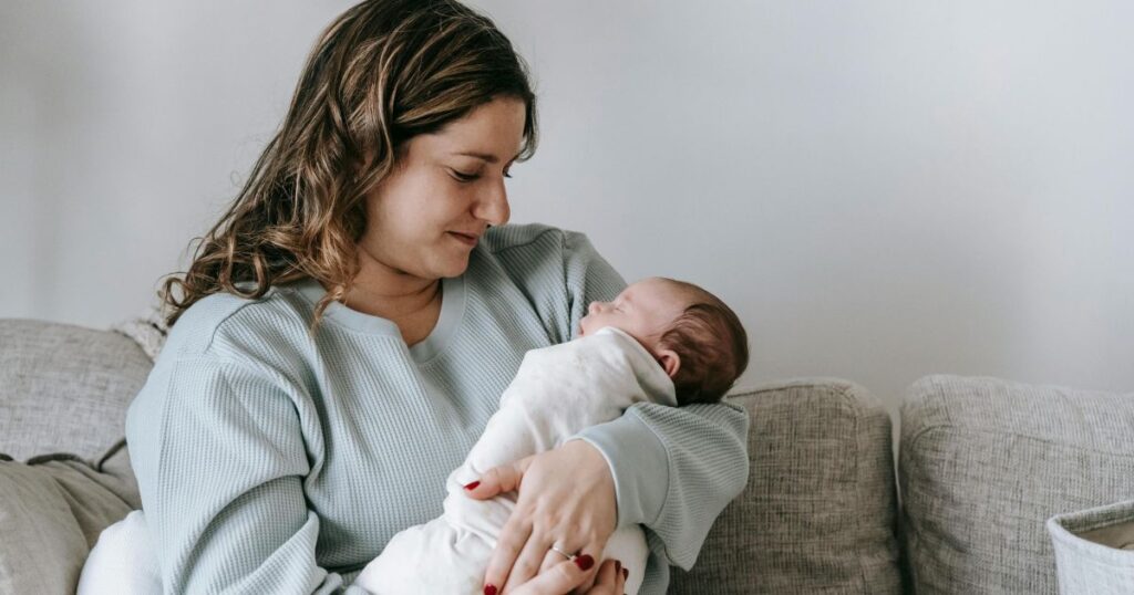 postpartum doula sitting on a grey couch holding a client's baby while baby sleeps