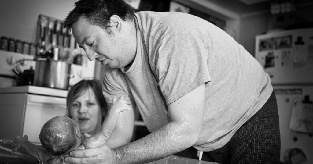 partner lifting newborn baby from the birthing tub at a home water birth in charlotte NC with a midwife observing