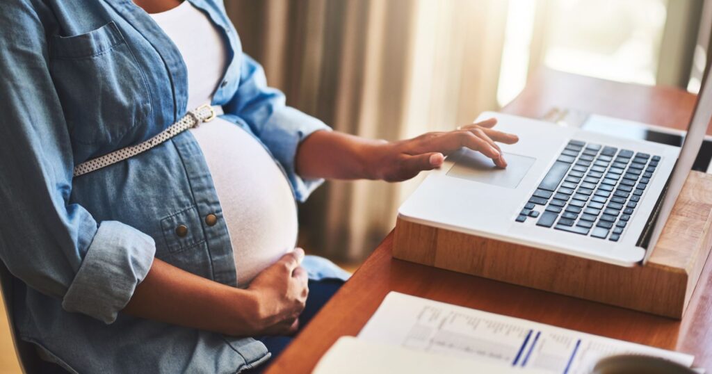pregnant person working on laptop at a table resting one hand on their belly