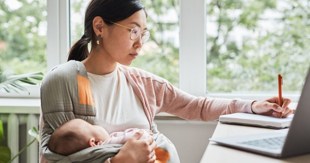 Mother holding a baby while working at a desk with a laptop and notebook