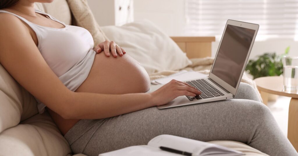 Woman sitting on a couch using a laptop while holding their belly