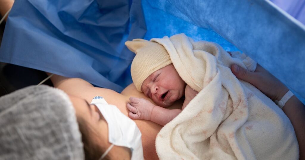 mother lying on a hospital bed wearing a mask and holding her crying newborn baby after giving birth