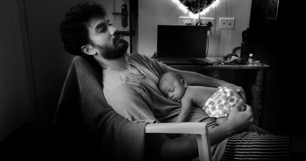 new father sitting on a chair at night, holding his sleeping newborn baby