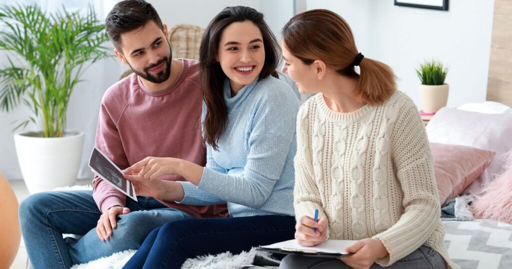 expecting parents holding an ultrasound smiling with a woman taking notes in the living room