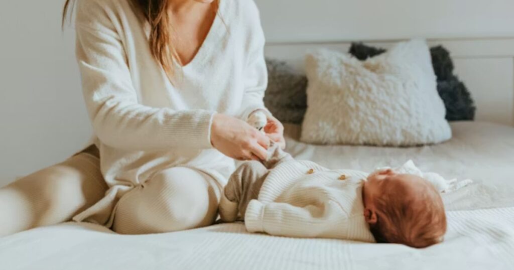 postpartum doula sitting on a bed with baby, putting on his sock