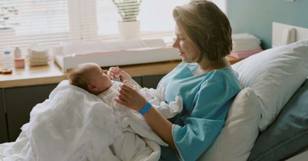 postpartum mother in a hospital gown sitting on a hospital bed smiling at her newborn baby in a white onesie