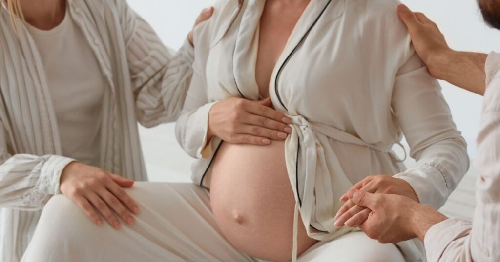 pregnant woman sitting with one hand on her belly being supported by partner and doula