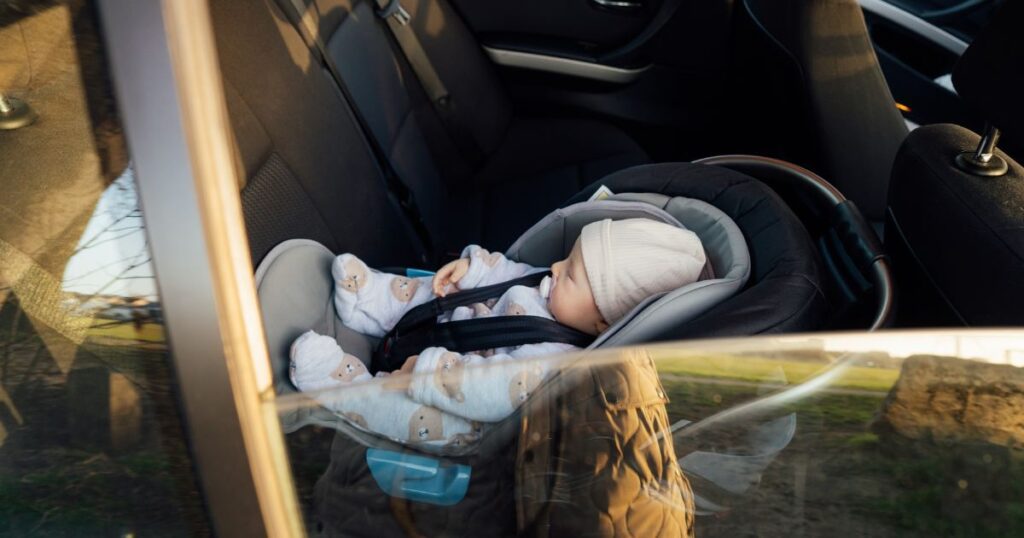 a photo taken from outside a car of a newborn baby wearing a onesie and peacefully sleeping while safely strapped inside a car seat