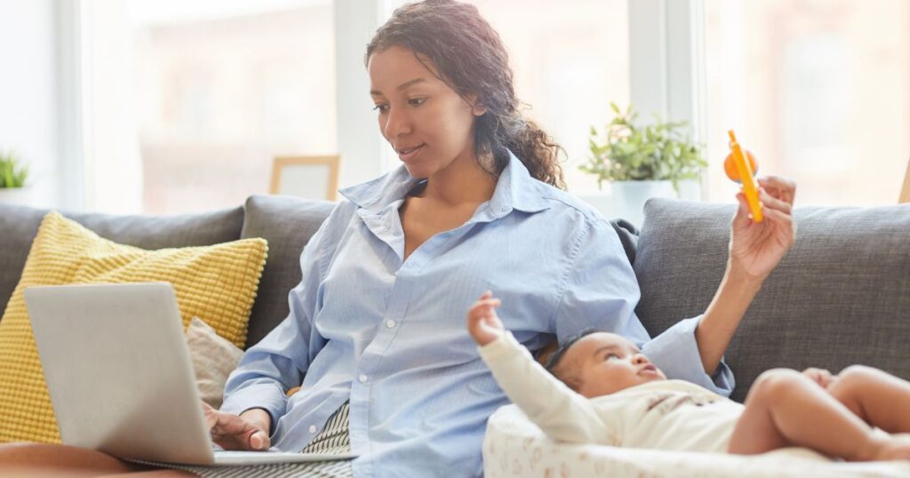 a mother sitting on her couch and scrolling on her laptop while holding her baby's toy and baby is looking up at it