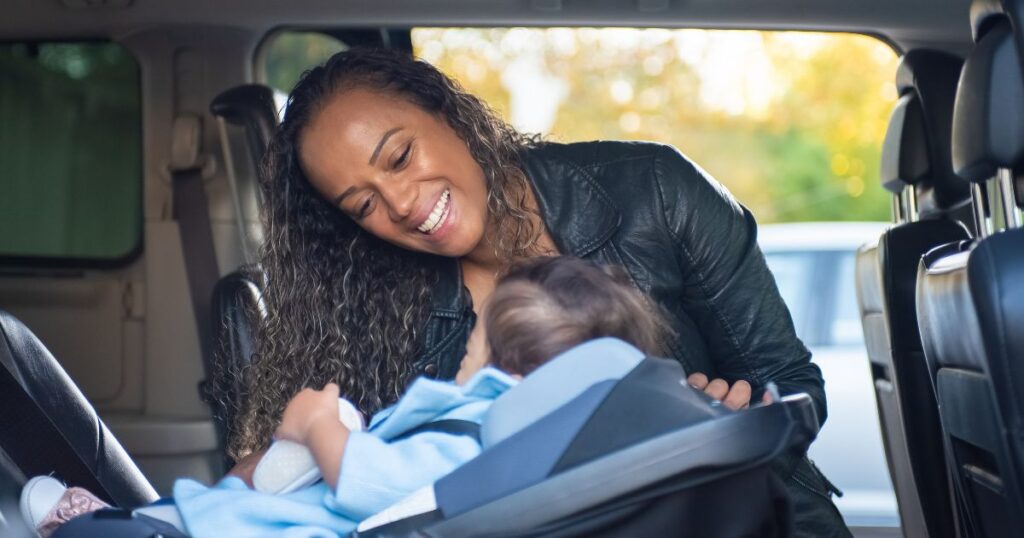 a smiling mother in a car, looking down at her baby who is safely buckled up in their rear-facing car seat