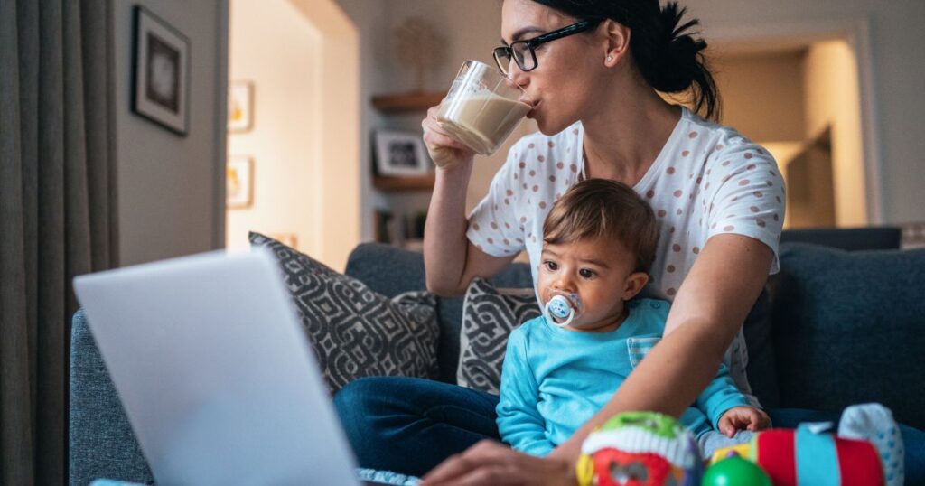a mother and her baby sitting comfortably on the couch as the mother drinks her coffee and checks the laptop while baby looks curiously at the screen