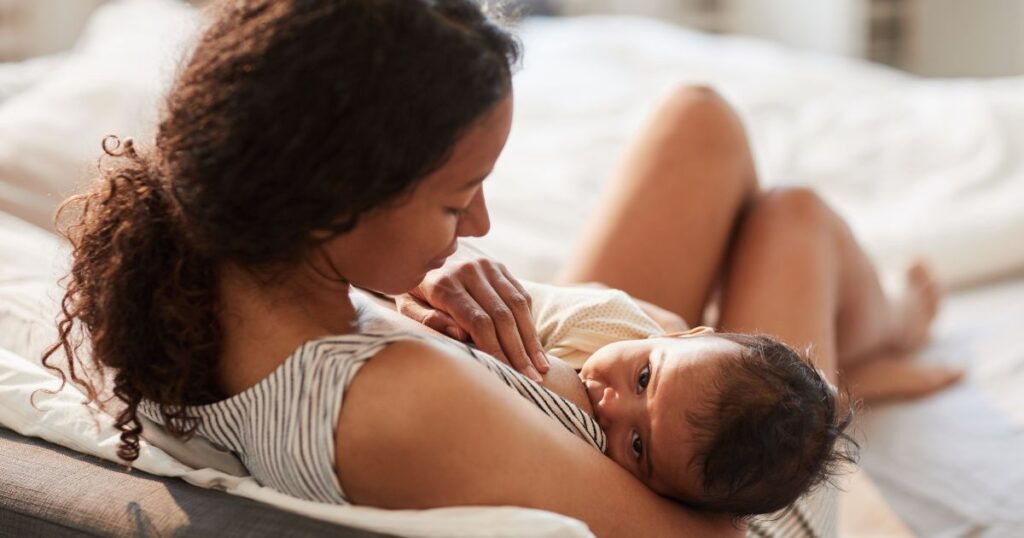 postpartum mother sitting on her bed, holding her baby while baby breastfeeds