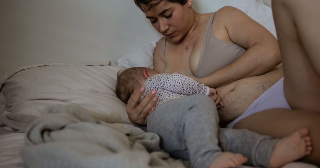 a newly postpartum mother in a nursing bra, lying on her bed while she holds her baby beside her while breastfeeding