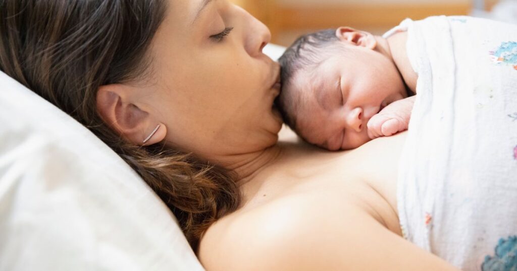 postpartum mother kissing and holding her sleeping newborn baby on her chest while baby sleeps