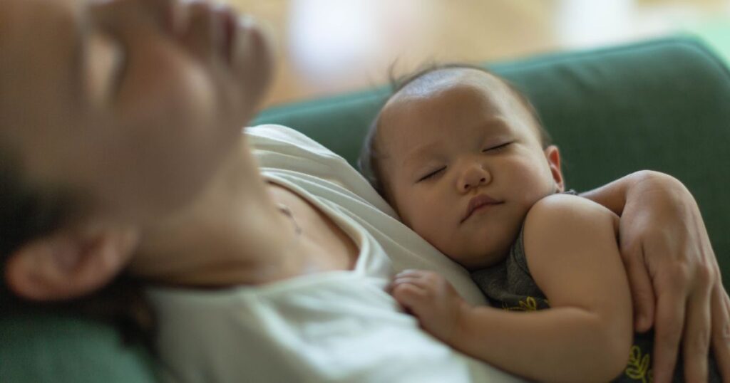 new dad lies asleep on couch while holding his sleeping baby in his arms