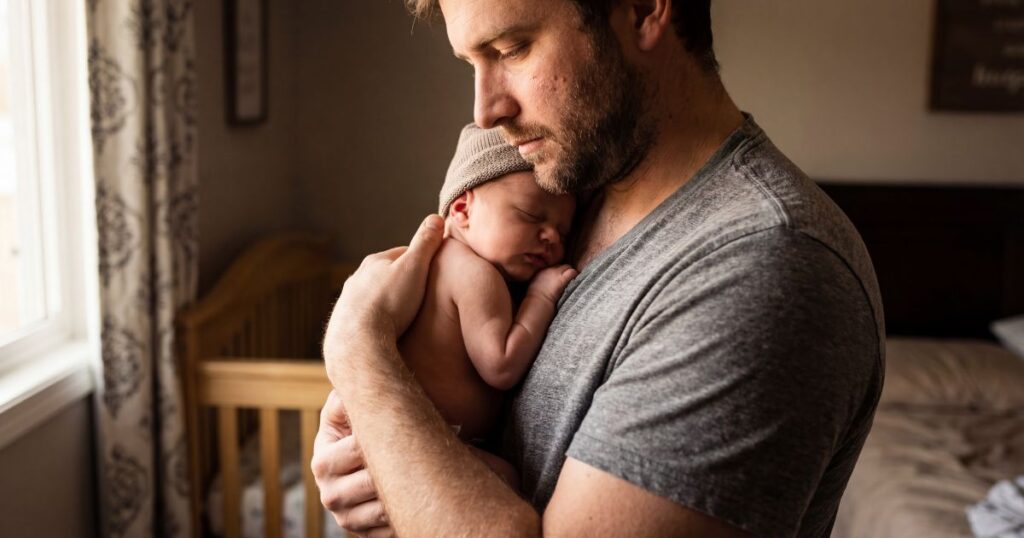 father holding his sleeping newborn baby close to his chest while looking down at the ground emotionally