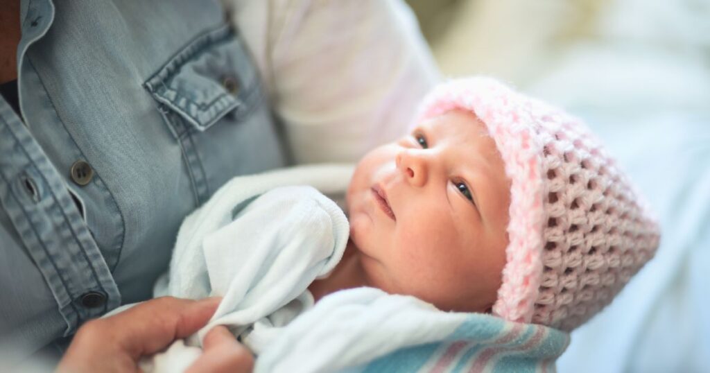 a close-up of a newborn baby wearing a pink knitted beanie, wrapped in a hospital blanket, looking up at the person holding them