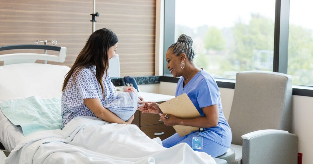 a newly postpartum mother sitting upright in a hospital bed, holding her newborn baby in her arms while she speaks with her doctor