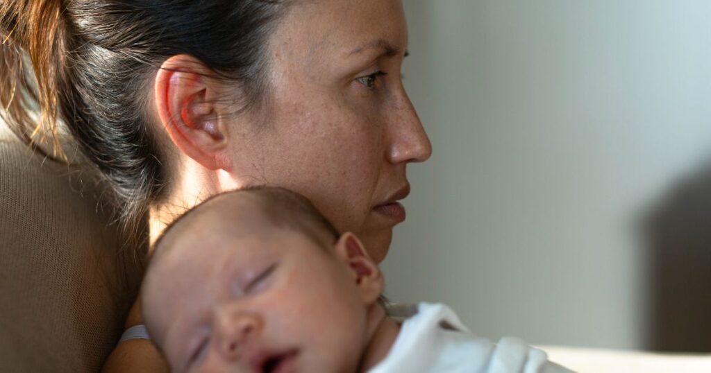 postpartum mother holding her sleeping newborn baby while she stares into the distance