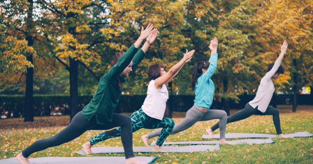 a group of women standing on yoga mats doing yoga outdoors on the grass in a public space